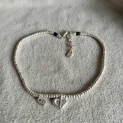 A silver beaded bracelet with two heart charms, one plain and one engraved with 'LOVE' and small clear stones, displayed on a textured light gray fabric background.