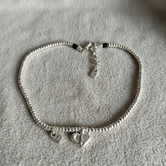 A silver beaded bracelet with two heart charms, one plain and one engraved with 'LOVE' and small clear stones, displayed on a textured light gray fabric background.