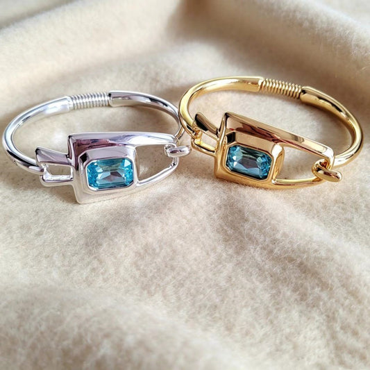 Two slim bangle rings with rectangular bezel-set light blue gemstones, one in polished silver and the other in polished yellow gold, displayed on a soft beige fabric background.