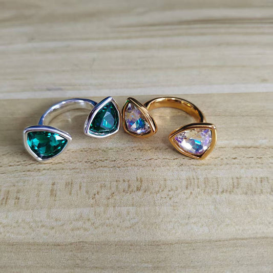 Four triangular bezel-set rings arranged in a row on a light wood surface. Two rings have green stones and two have iridescent blue-purple stones, with one ring in silver and three in gold.