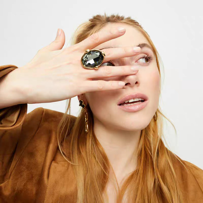 A close-up lifestyle shot of a woman with blonde hair wearing a large gold ring with a dark oval gemstone, holding her hand in front of her eye.