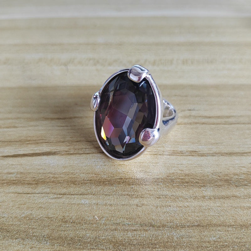 A silver ring featuring a large, faceted oval gemstone with a dark purple and pink iridescent sheen, resting on a light wood grain surface.
