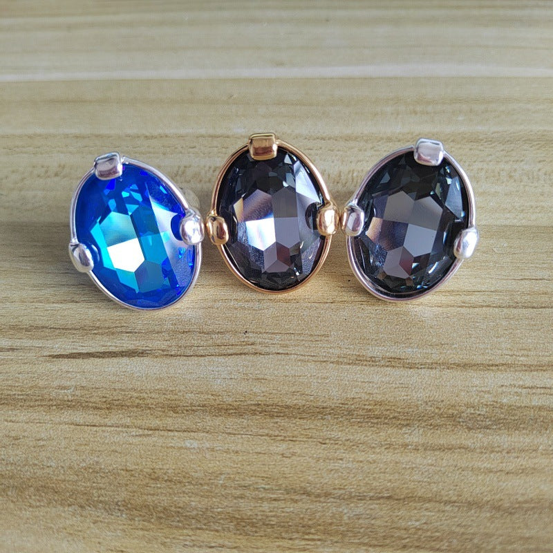 Three faceted oval gemstone stud earrings in silver and rose gold settings, displayed on a light wood background. The left earring features a vibrant blue stone, while the center and right earring features a dark smoky gray stone.