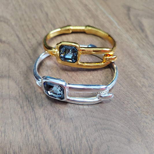 Two slim open bangle rings with rectangular blue gemstones, one in polished gold and one in polished silver, displayed on a wooden surface.