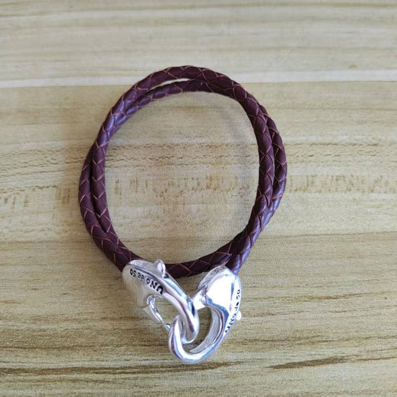 A dark purple braided leather bracelet with a silver heart-shaped clasp, displayed on a light wood grain surface.