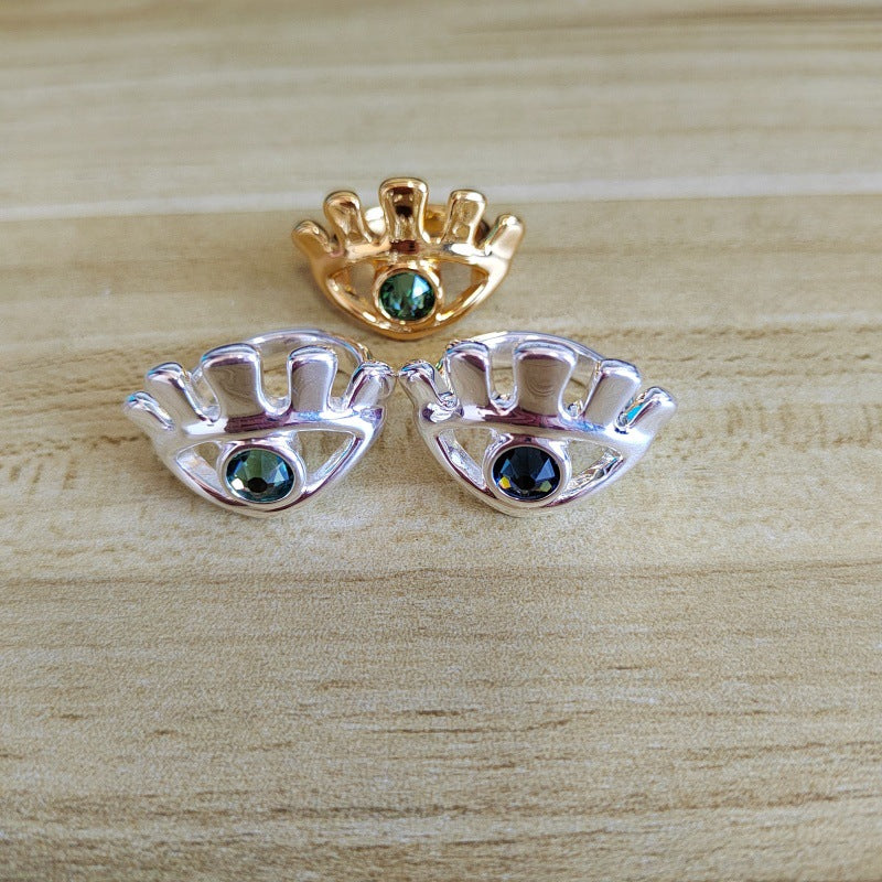 Three small crown-shaped jewelry pieces with iridescent green gemstones, shown on a light wood surface: one gold-toned piece and two silver-toned pieces.