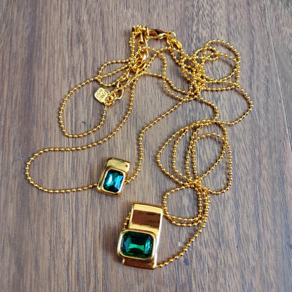 A gold chain necklace with two rectangular pendants featuring large green faceted crystals, displayed on a dark wood background.