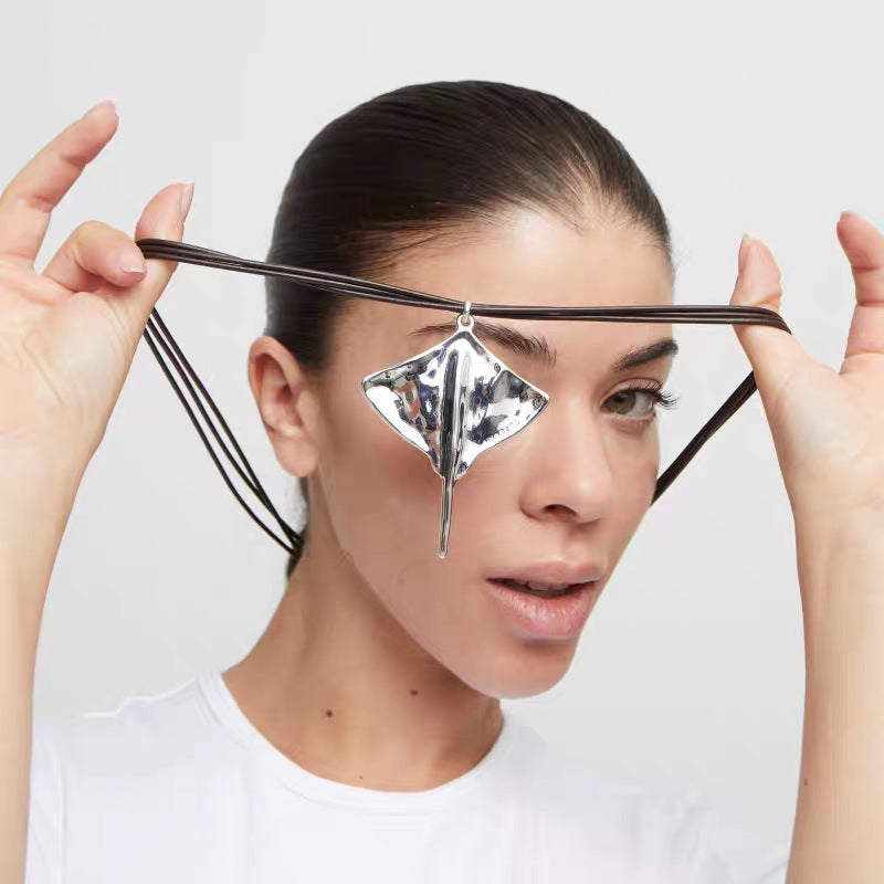 A woman wearing a white t-shirt holds a black elastic headband with a large, faceted silver geometric pendant across her forehead against a light gray background.