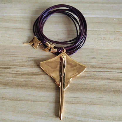 A gold-toned pendant necklace shaped like a manta ray, paired with a multi-strand wrap bracelet in dark purple and black cords, displayed on a light wood grain surface.