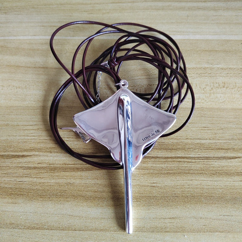 A silver pendant necklace featuring a stylized fish or eel design with a vertical bar, resting on a coiled dark brown leather cord, set against a light wood grain background.