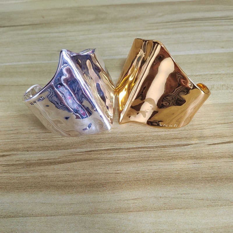 Two sculptural, wavy metal rings in polished silver and rose gold finishes, displayed side-by-side on a light wood grain surface.