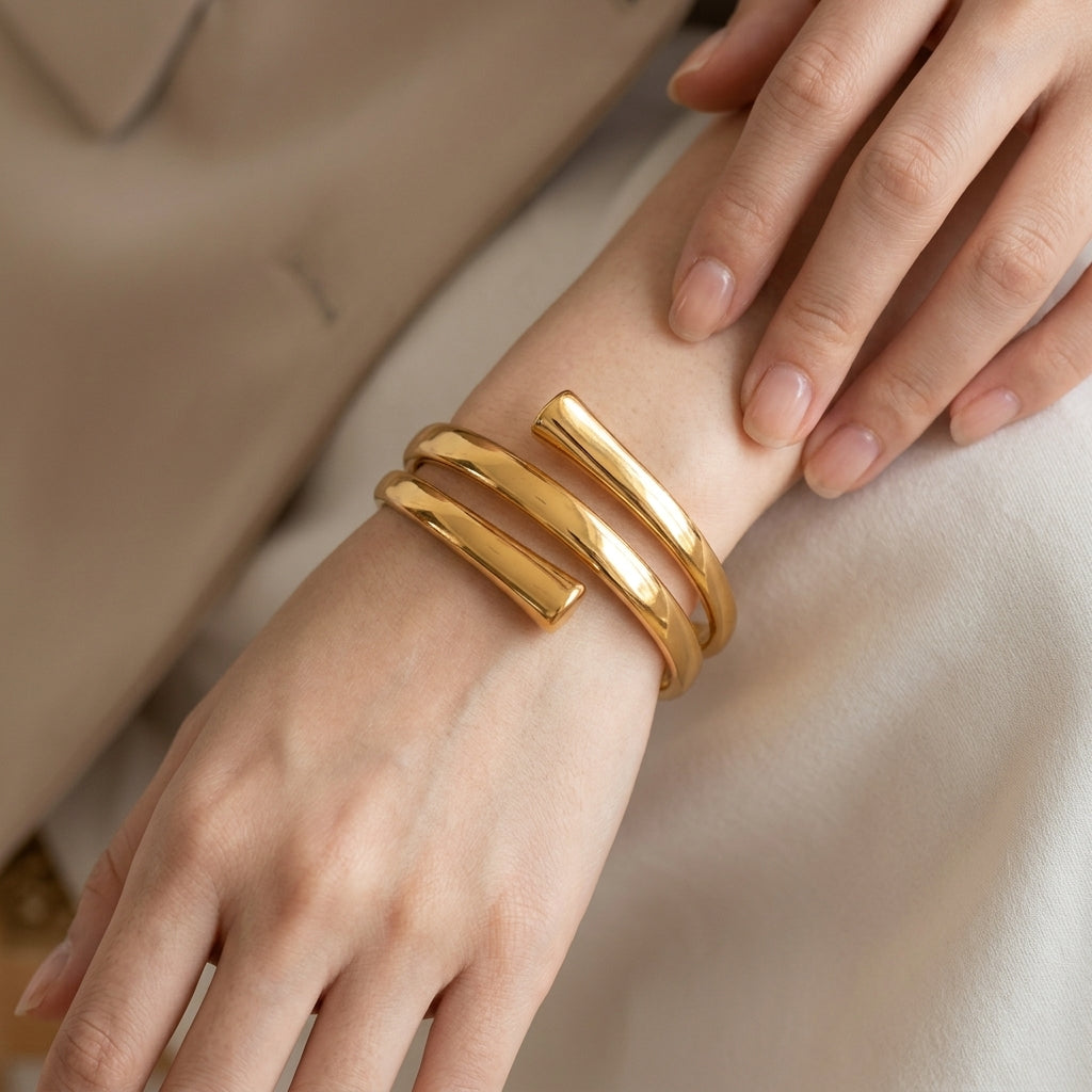 A close-up of a person's wrist wearing a polished gold open cuff bracelet, with their hands resting near their lap against a soft beige and cream background.