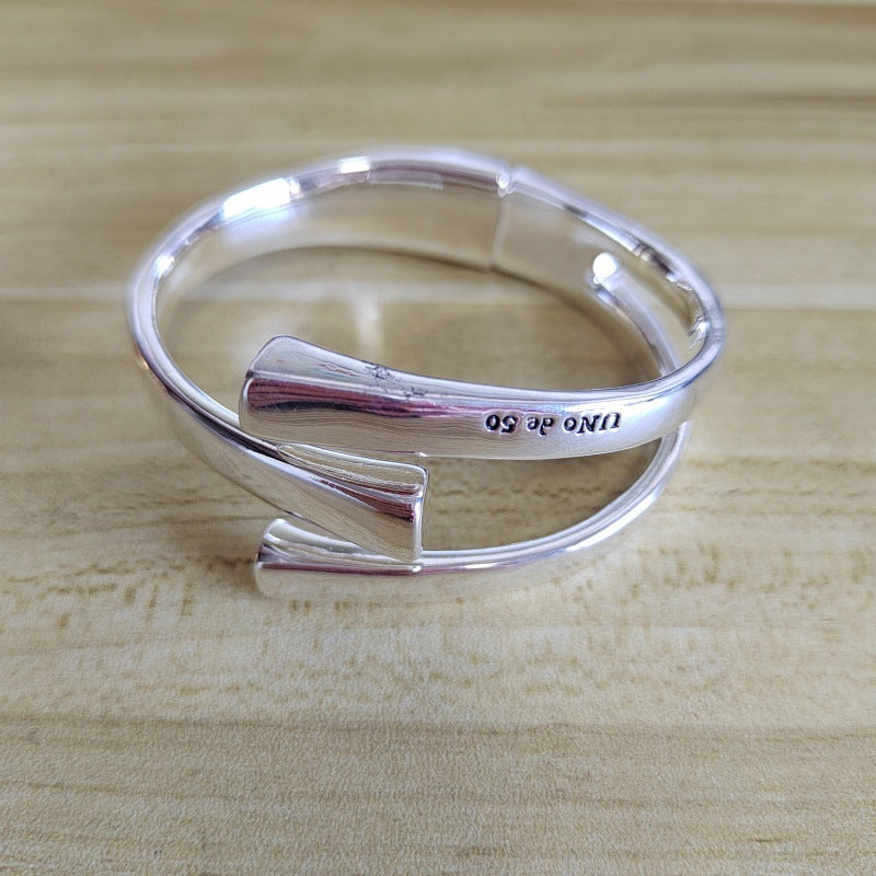 A polished silver open cuff bangle bracelet with a polished finish and engraved text 'UNO DE 50' resting on a light wood surface.
