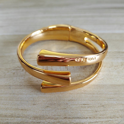 A polished gold-tone wrap cuff bracelet with a sleek, modern design, resting on a light beige wood-textured surface. The brand name 'OROMI' is engraved on the side.