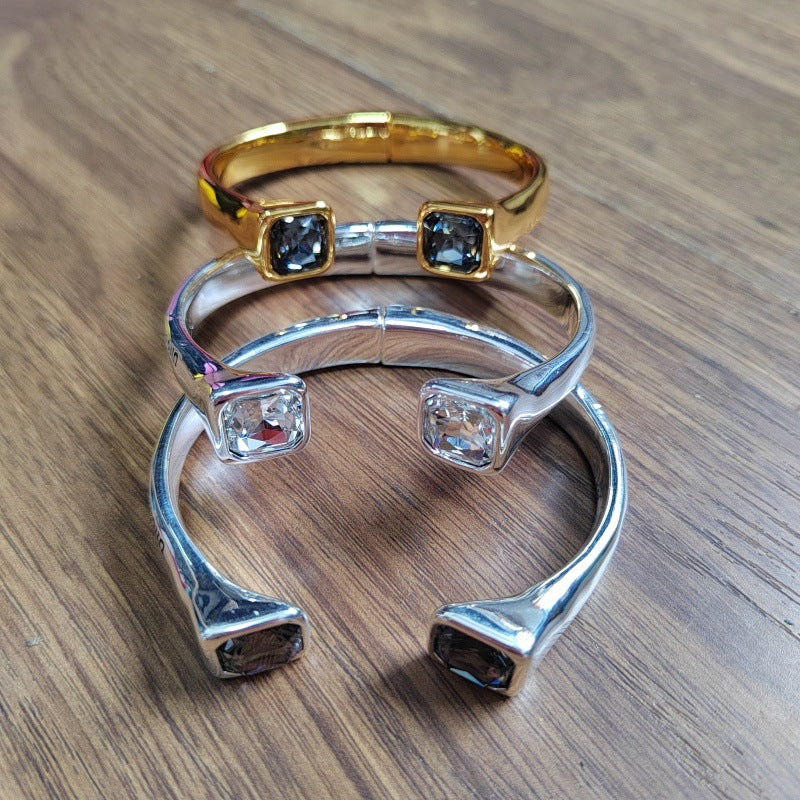 Three open cuff bracelets stacked on a wooden surface: one gold-tone with dark blue stones, one silver-tone with clear stones, and one silver-tone with dark brown stones.