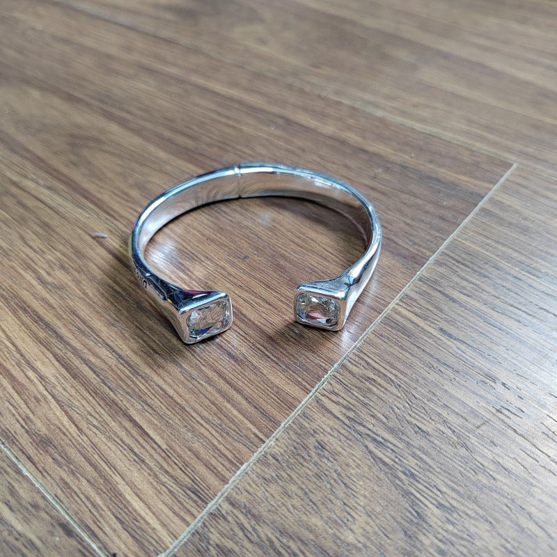 A silver-toned open cuff bracelet with square bezel-set clear stones on each end, photographed on a brown wood floor.