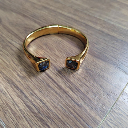 A gold-toned open cuff ring with square-cut dark blue gemstones on each end, photographed on a brown wood-grain surface.