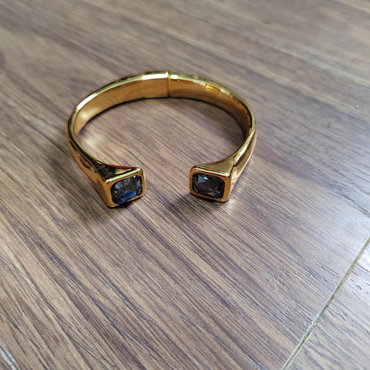 A gold-toned open cuff ring with square-cut dark blue gemstones on each end, photographed on a brown wood-grain surface.