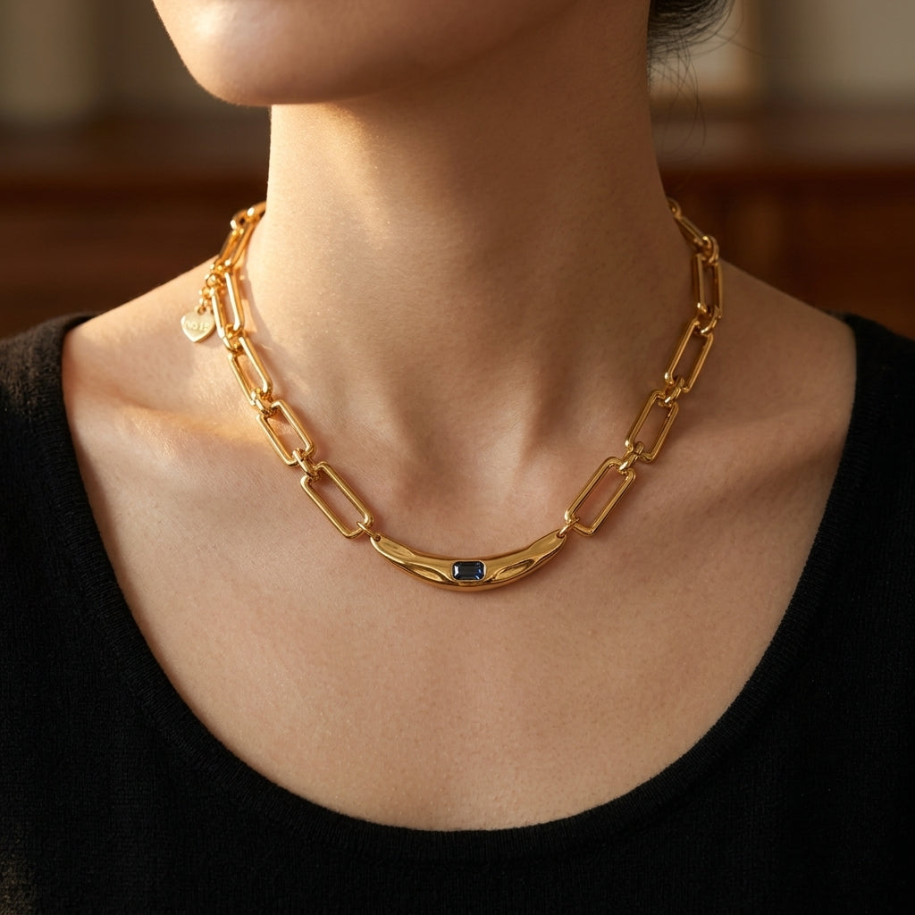 A close-up of a model wearing a gold chain necklace with a central dark blue stone and a small gold heart charm, paired with a black top.