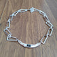 A silver geometric link bracelet with a curved center bar and a small dark gemstone, displayed on a brown wooden surface.