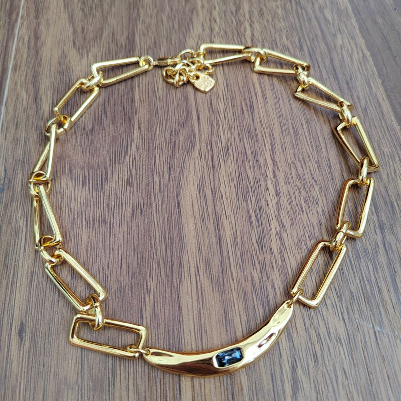 A gold chain necklace with a curved bar featuring a small dark blue stone, displayed on a brown wooden surface.