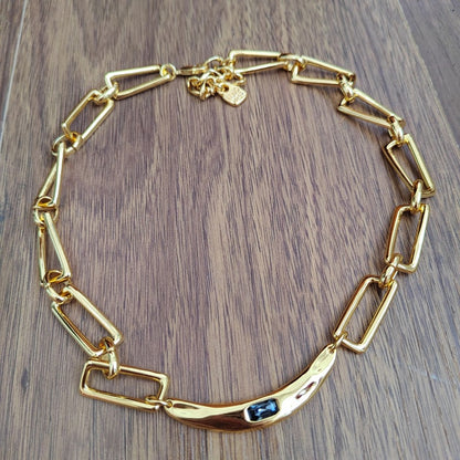A gold chain necklace with a curved bar featuring a small dark blue stone, displayed on a brown wooden surface.