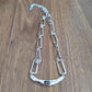 A silver-toned chain necklace with elongated rectangular links and a curved bar pendant featuring a small dark inlay, displayed on a brown wood surface.
