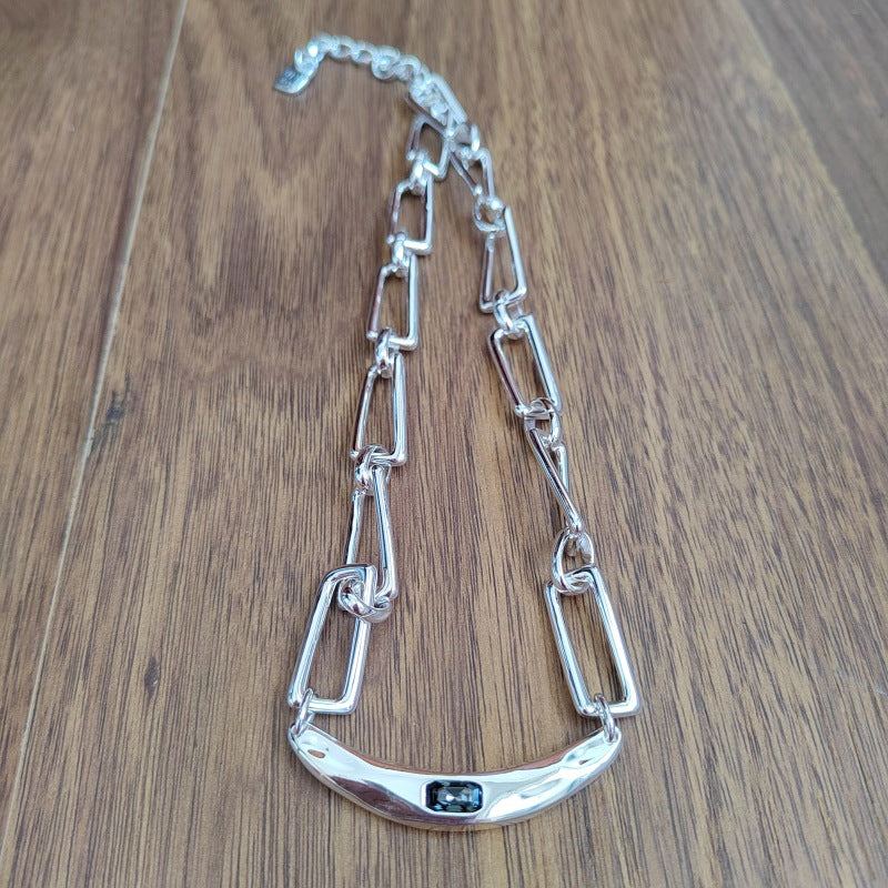A silver-toned chain necklace with elongated rectangular links and a curved bar pendant featuring a small dark inlay, displayed on a brown wood surface.