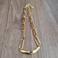 A gold-toned link chain necklace with a curved bar pendant featuring a small dark blue stone, displayed on a brown wooden surface.