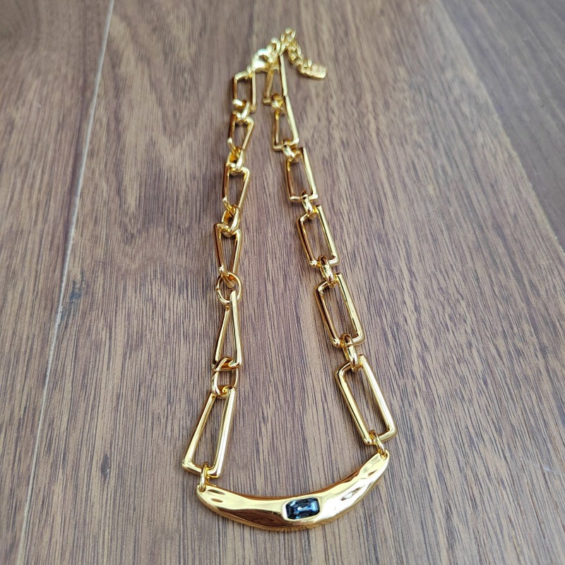A gold-toned link chain necklace with a curved bar pendant featuring a small dark blue stone, displayed on a brown wooden surface.