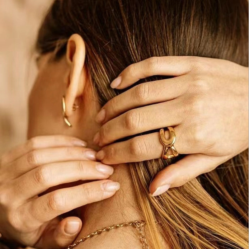 A close-up lifestyle shot of a woman wearing a gold ring with a bow-like design on her finger, with her hands touching her hair and ear.