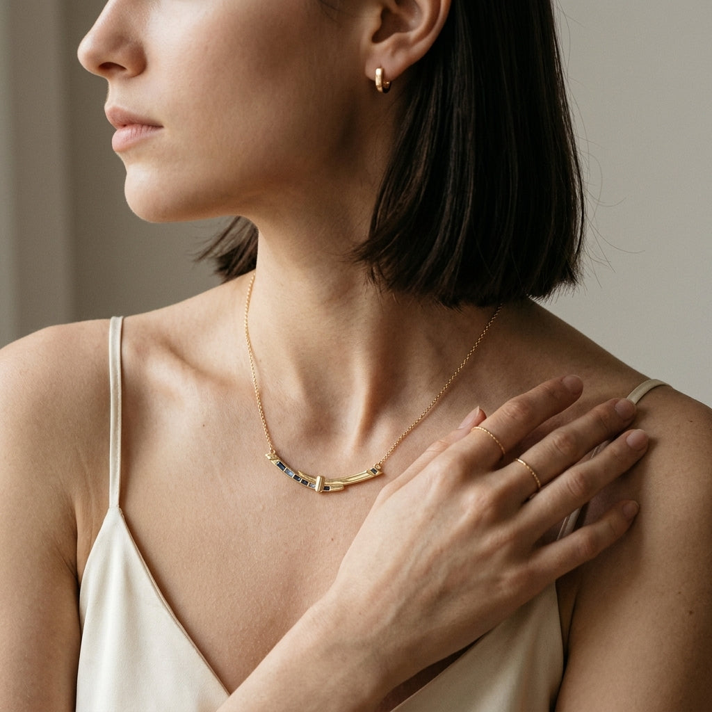 A close-up lifestyle shot of a woman wearing a delicate gold necklace with a central bar featuring small blue and pink gemstones, paired with a matching gold hoop earring and ring.