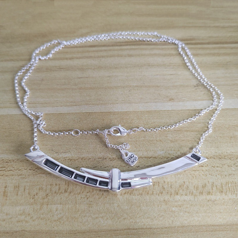 A silver-toned necklace featuring a curved bar pendant with rectangular dark inlay details, displayed on a light wood grain surface.