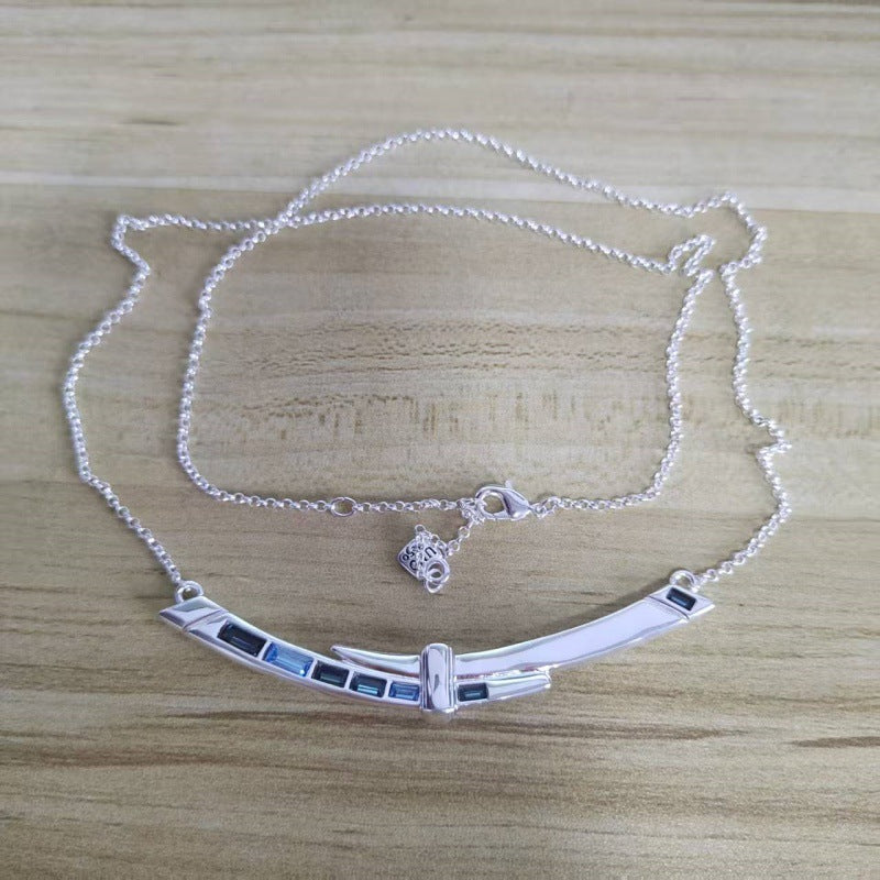 A silver-toned necklace with a curved bar pendant featuring inlaid dark blue rectangular stones, displayed on a light wood grain surface.