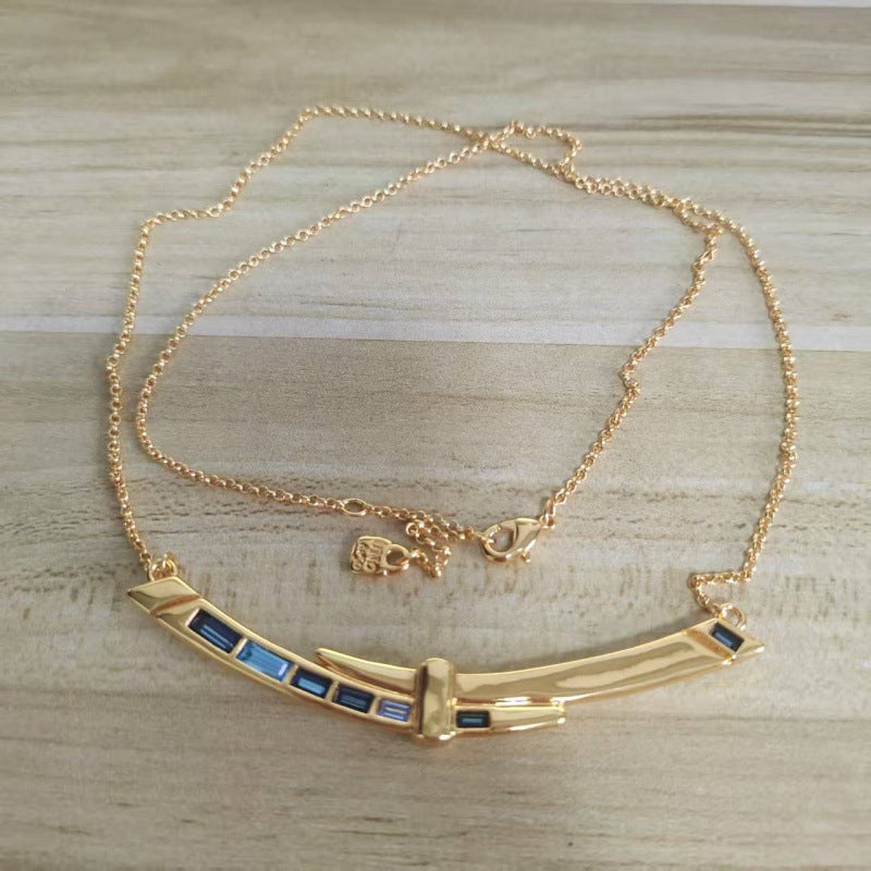 A gold-toned necklace with a curved bar pendant featuring small blue rectangular inlays, displayed on a light wood surface.