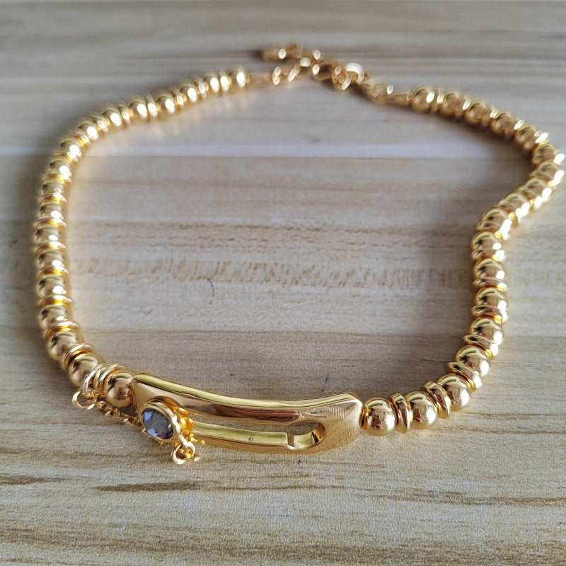 A gold-plated beaded bracelet with a curved bar centerpiece and a small oval blue stone, displayed on a light wood surface.