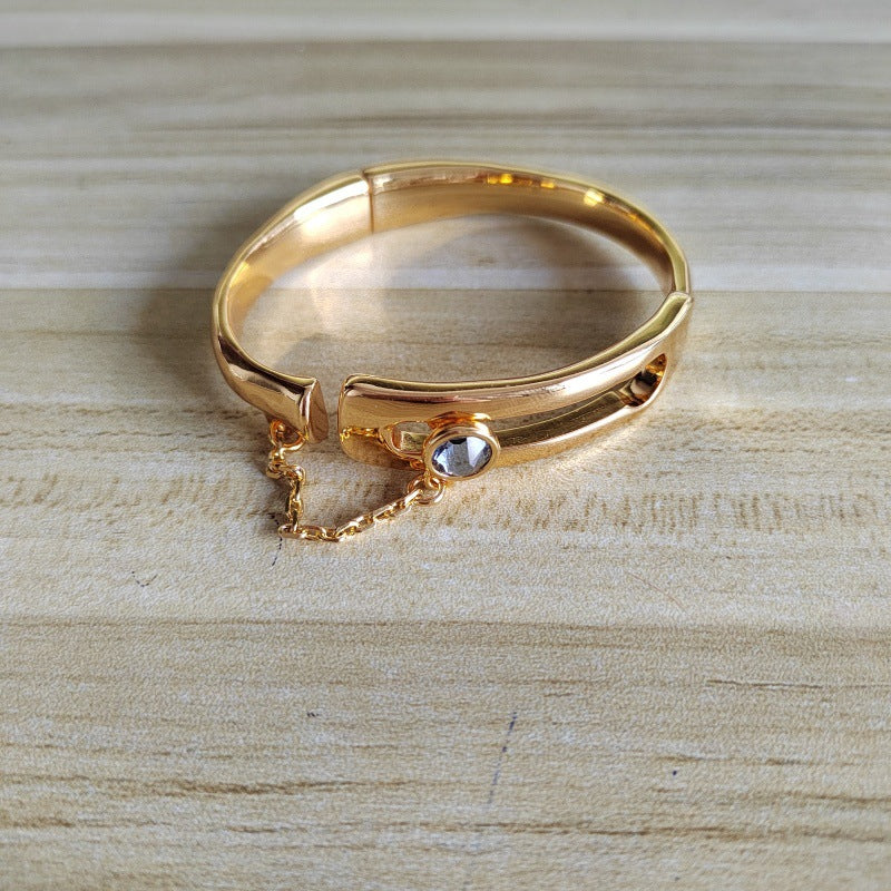 A gold bangle bracelet with a small clear rhinestone and an adjustable safety chain, displayed on a light wood grain surface.