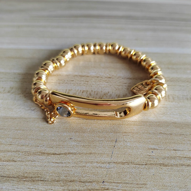 A gold-plated beaded bracelet with a central bar clasp and a small clear stone, displayed on a light wood grain surface.