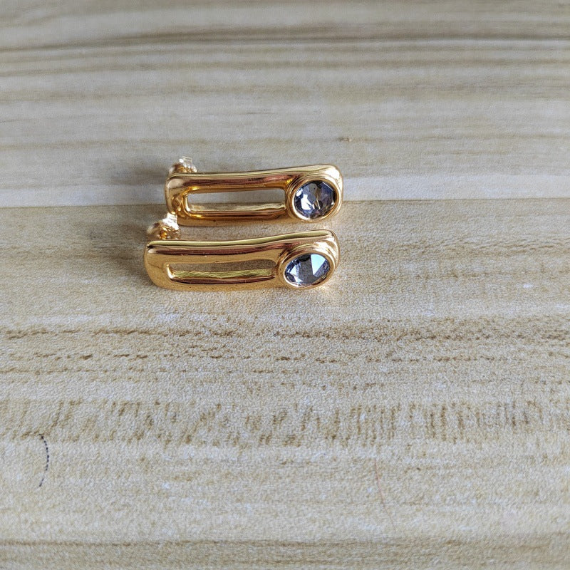 A pair of gold-toned bar earrings with a small clear teardrop gemstone on each, displayed on a light wood grain surface.