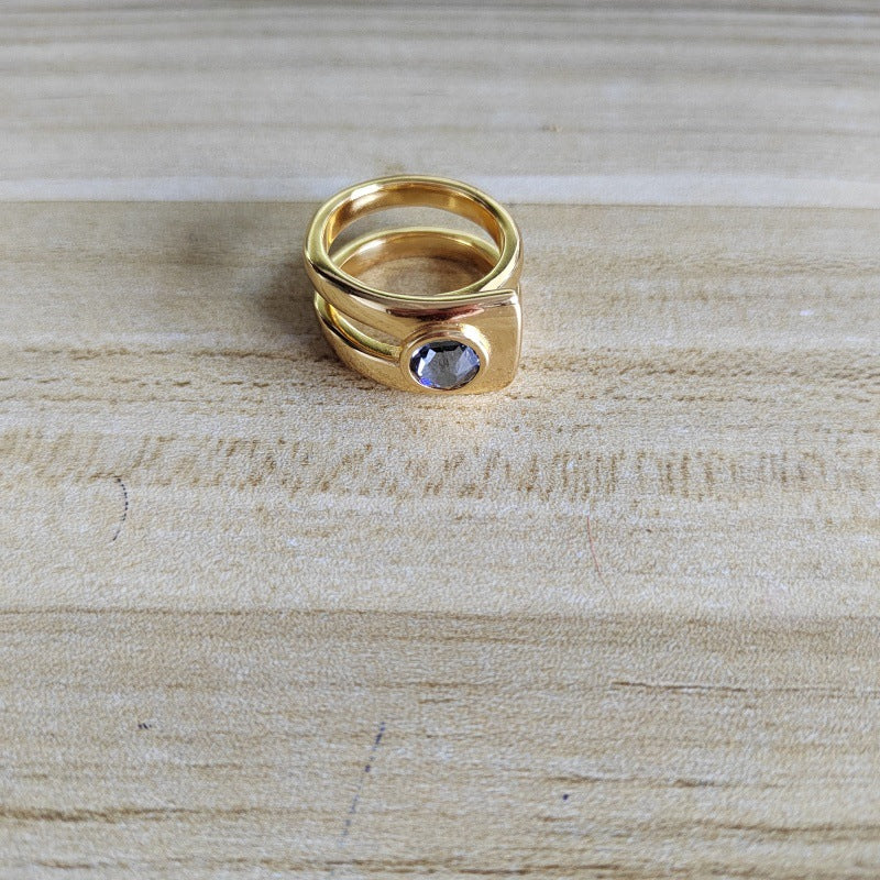 A gold-plated ring with a square bezel setting holding a round, clear gemstone, resting on a light wood grain surface.