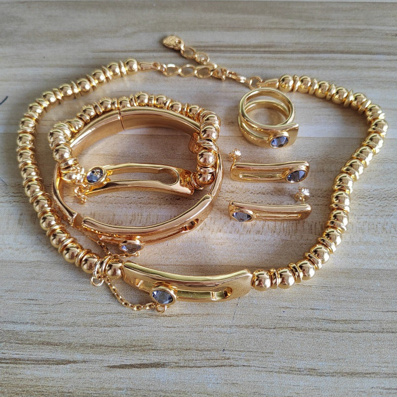 A gold-toned jewelry set featuring a beaded chain necklace, a matching bangle bracelet, a ring, and slim bar earrings, each accented with small blue gemstones, displayed on a light wood surface.