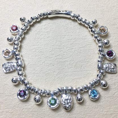 A sterling silver charm bracelet with multiple dangling charms, including colorful gemstone beads and textured silver charms, displayed on a textured beige surface. The clasp is engraved with 'UNIQUE SO'.