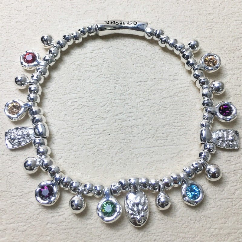 A sterling silver charm bracelet with multiple dangling charms, including colorful gemstone beads and textured silver charms, displayed on a textured beige surface. The clasp is engraved with 'UNIQUE SO'.