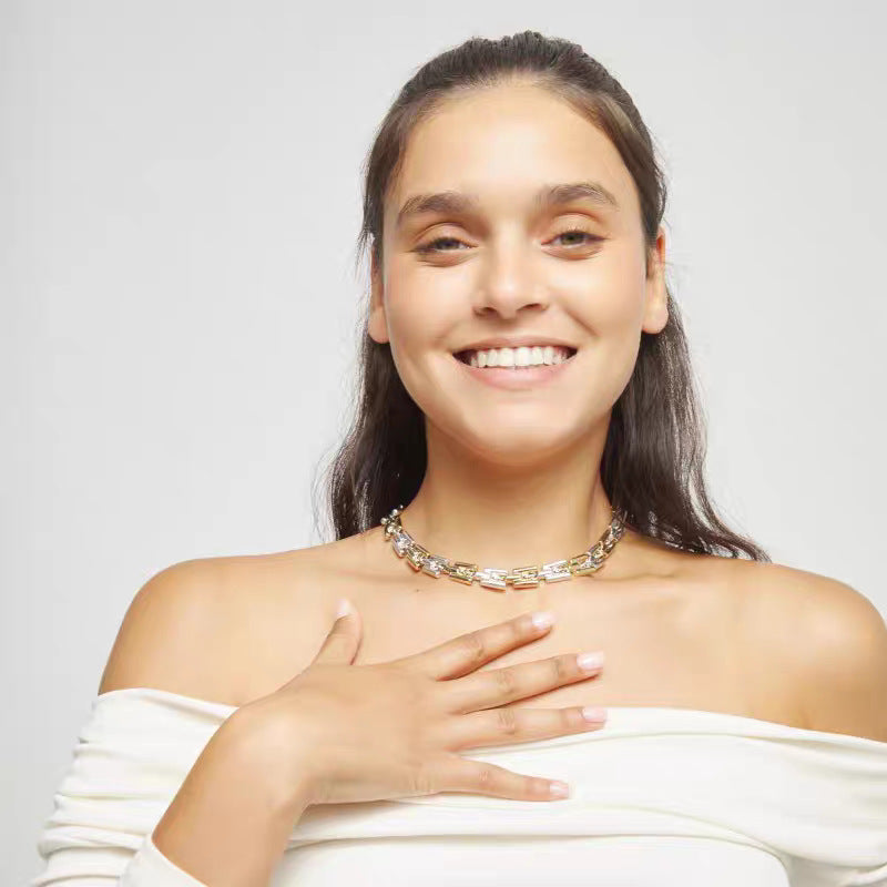 A smiling woman with dark hair pulled back wears a chunky gold chain necklace with clear stones and a white off-the-shoulder top against a light gray background.