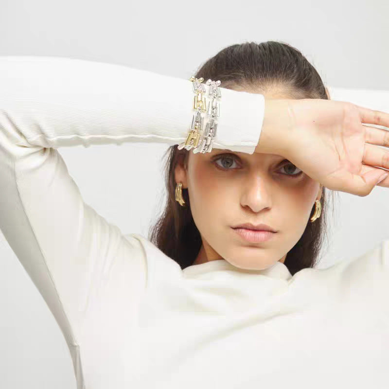 A woman wearing a white ribbed long-sleeve top poses with her arm raised, showcasing a stack of chunky silver and gold chain bracelets on her wrist. She is also wearing small gold hoop earrings against a clean, light gray studio background.