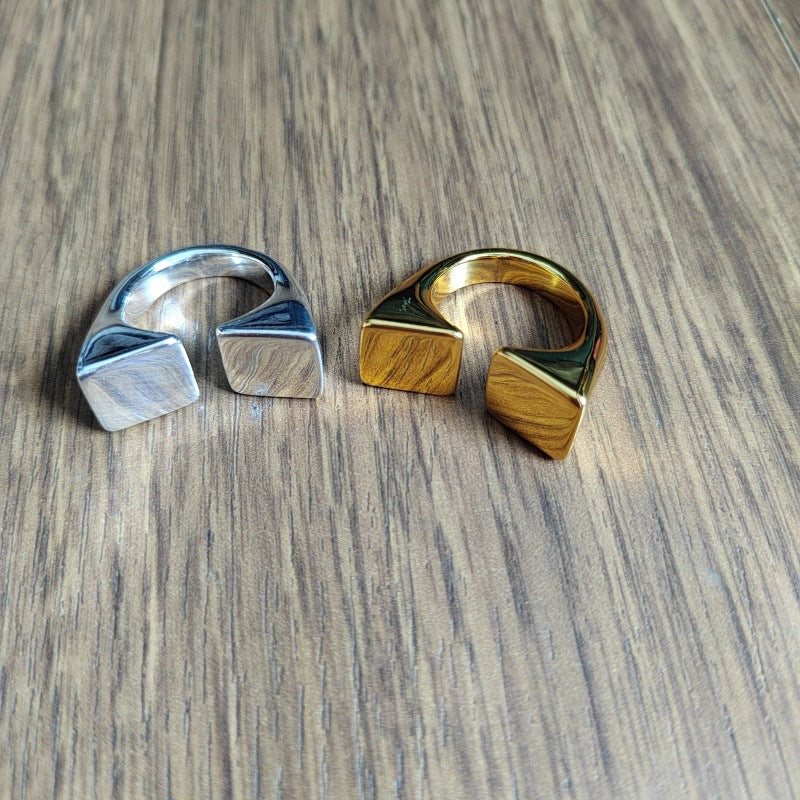 Two geometric open cuff rings with square ends, one in silver-toned metal and one in gold-toned metal, displayed on a wood grain surface.