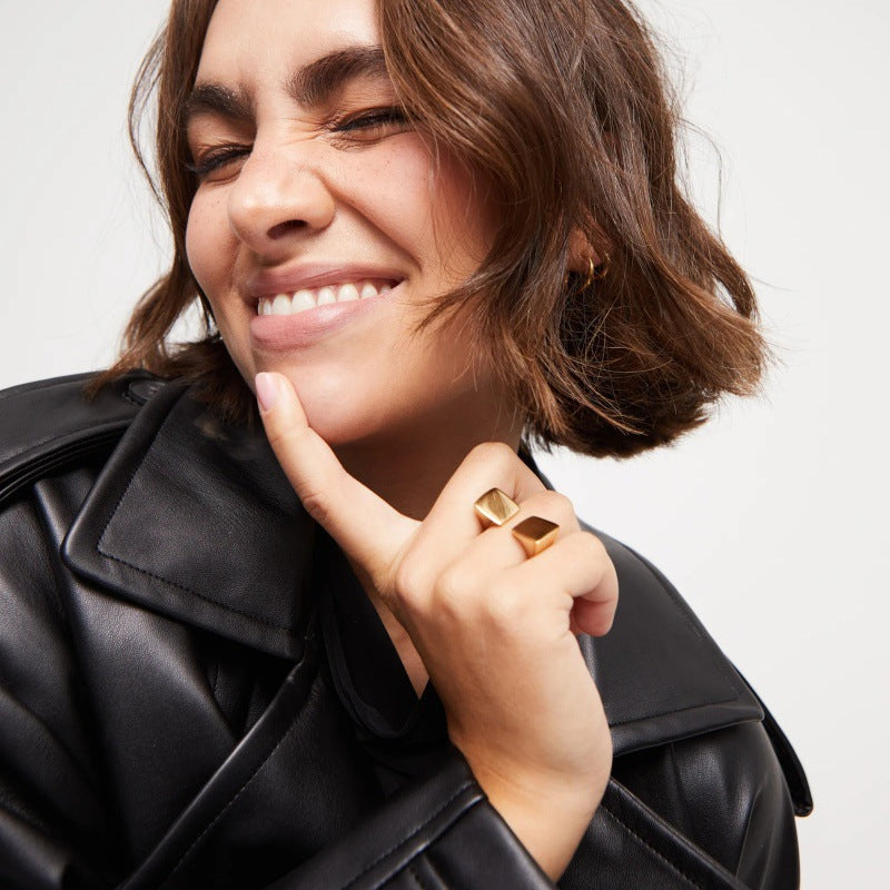 A close-up lifestyle shot of a woman with short brown hair wearing a chunky gold geometric ring and a black leather jacket, posing with her hand near her face against a light gray background.