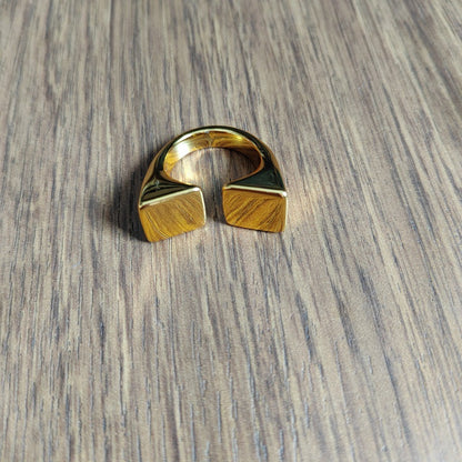 A gold-toned geometric ring with a split square design, resting on a gray wood-grain surface.
