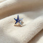 A silver starfish-shaped stud earring with a glossy blue enamel center, resting on a soft, cream-colored textured fabric.