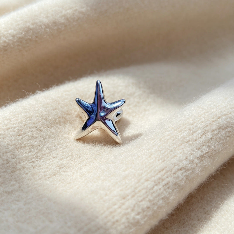 A silver starfish-shaped stud earring with a glossy blue enamel center, resting on a soft, cream-colored textured fabric.
